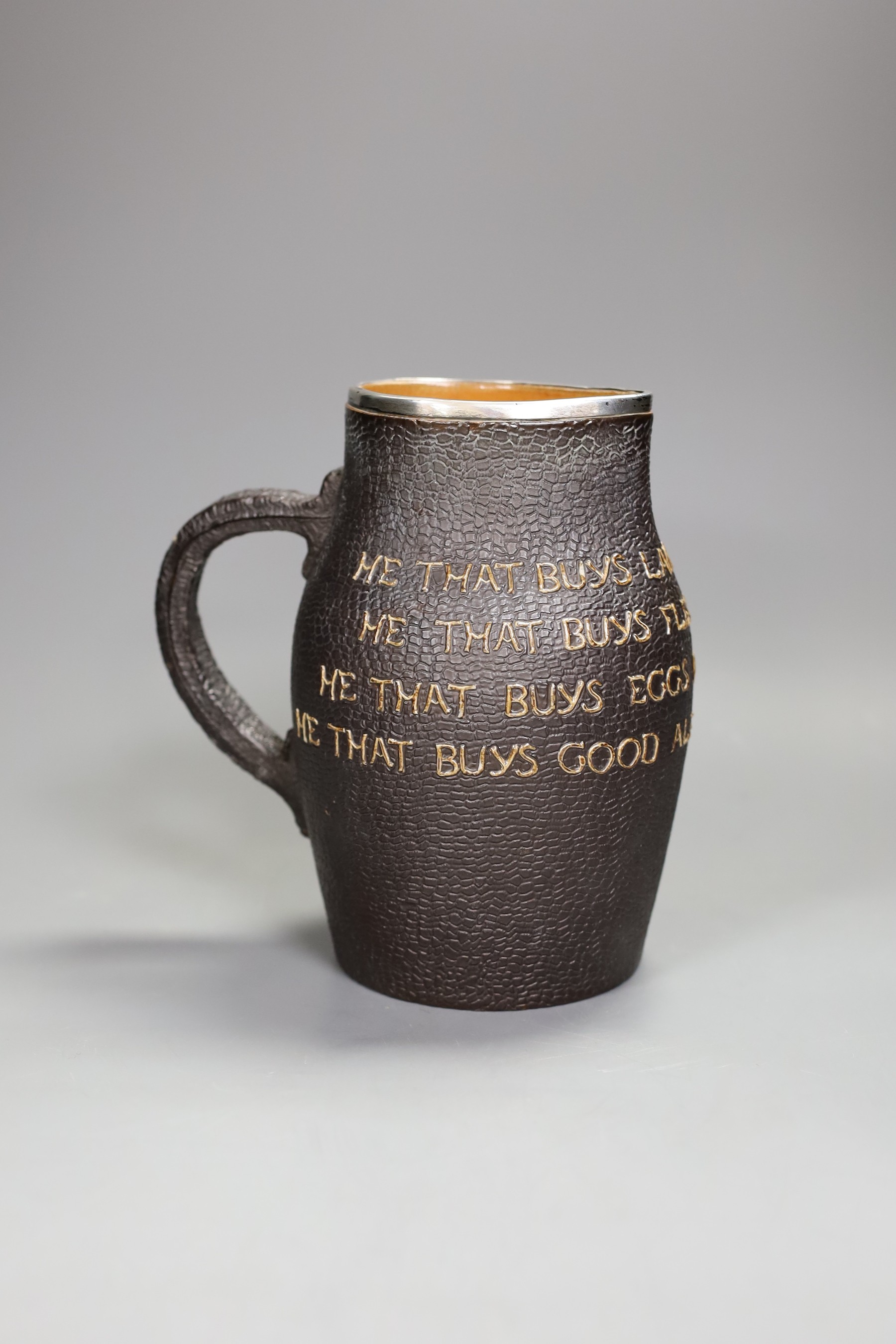 A Doulton Lambert leather-effect jug, ‘He Who Buys Good Ale Buys Nothing Else’, with silver mount, 19cm tall